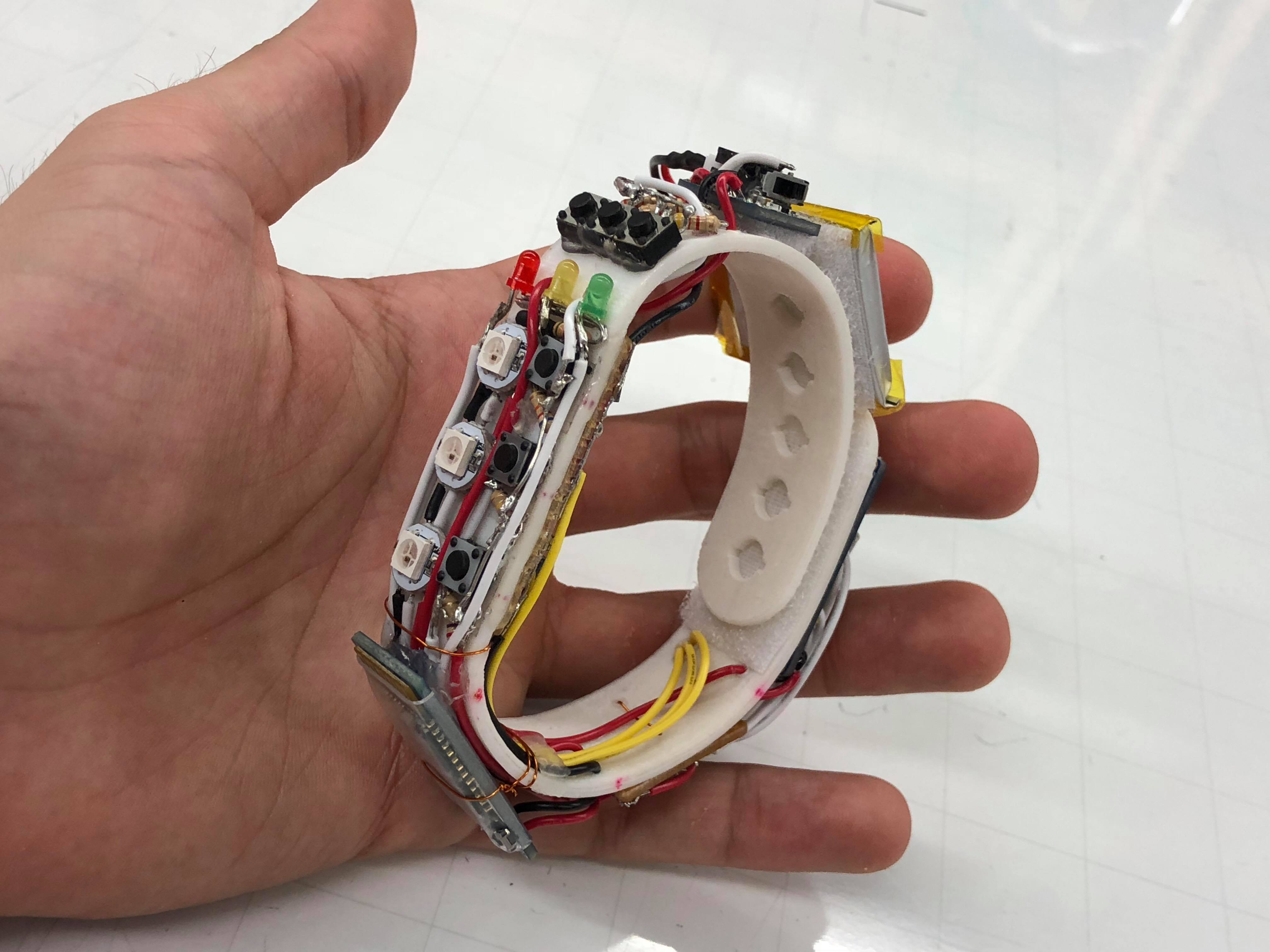Photo depicts a prototype of an electronic wrist band in the hand of a Siempre team member. There are exposed wires, buttons, lights and other components. The band is white, curved and appears to be 3D-printed.